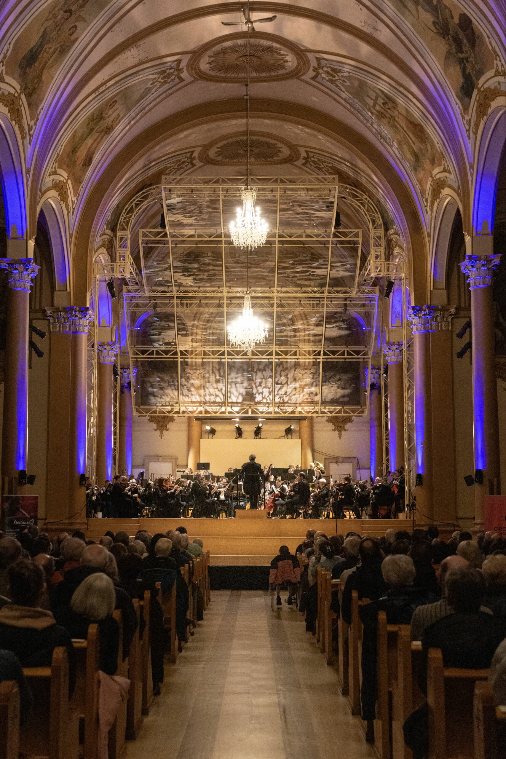 An orchestra performs on stage in a grand hall with ornate arched ceilings, chandeliers, and purple lighting, as an audience sits in rows facing the musicians.