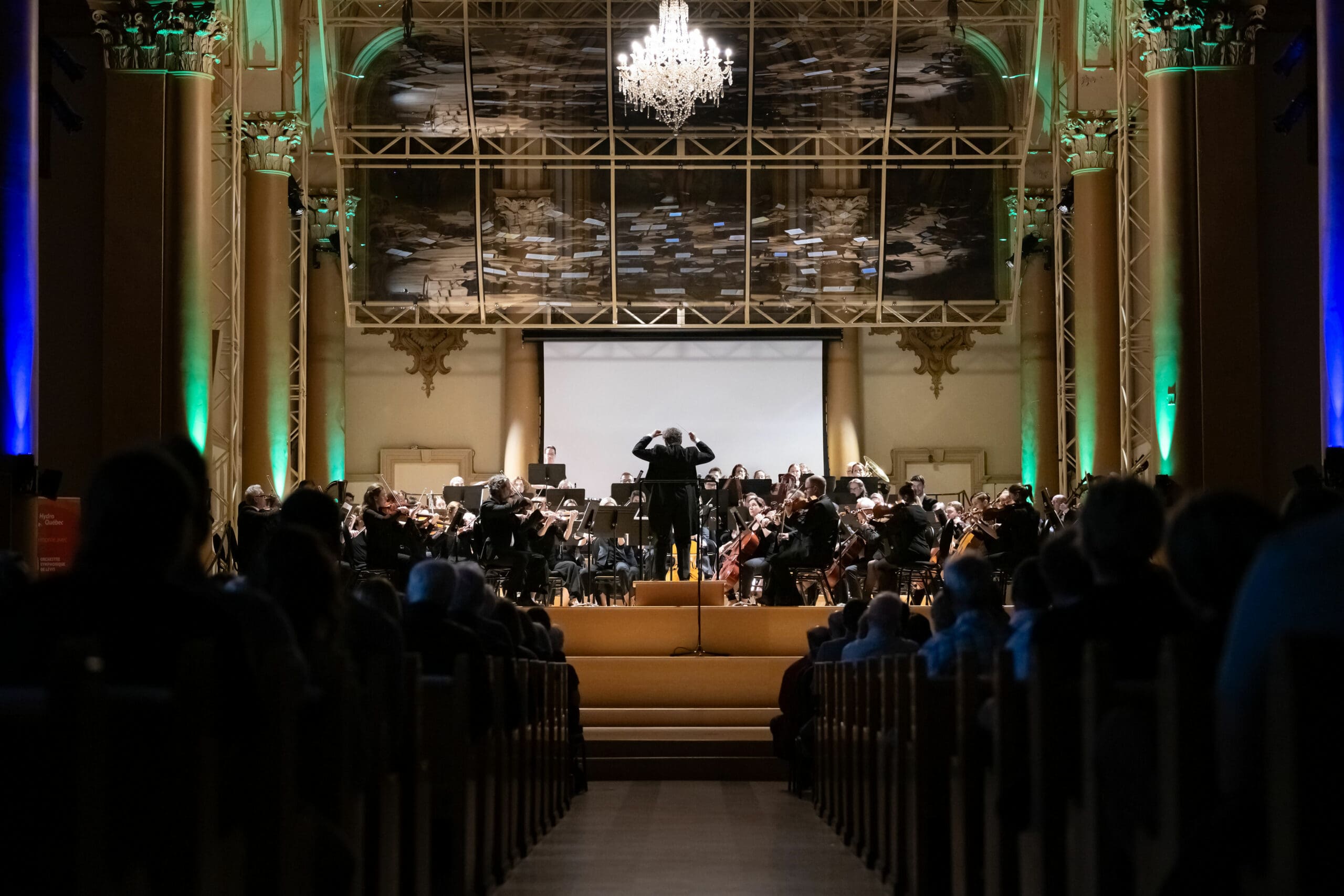 A conductor leads an orchestra performing on a stage in an ornate concert hall, viewed from the back of the audience. The hall is decorated with columns, a large chandelier, and a painted ceiling.