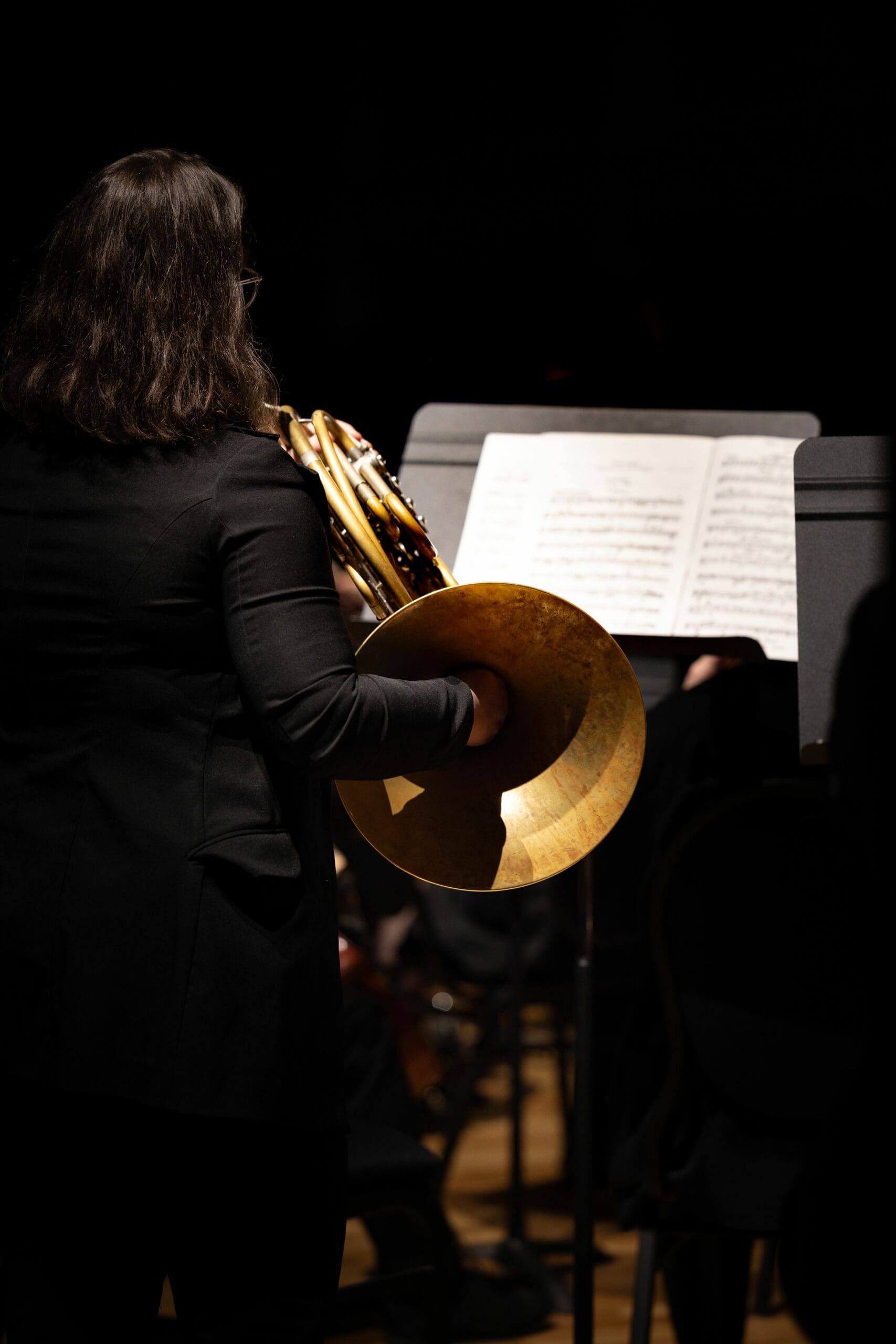 A musician with long dark hair, dressed in black, plays a French horn while standing in front of a music stand with sheet music, illuminated by stage lighting against a dark background.