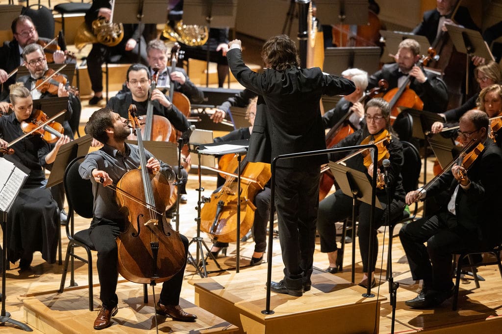 A conductor leads an orchestra onstage, with musicians playing various string instruments, including a prominent cellist in the foreground. The musicians are focused on their music and the conductor’s direction.