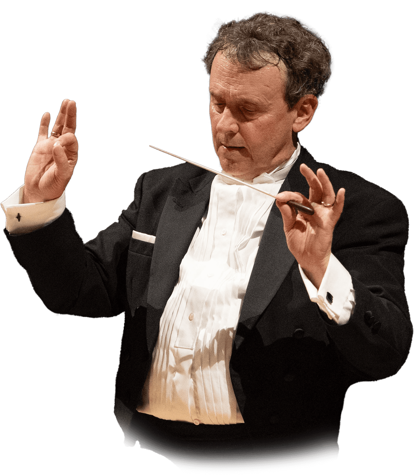A man in formal attire conducts music, holding a baton and gesturing with his hands. He appears focused and is dressed in a black tuxedo with a white shirt.