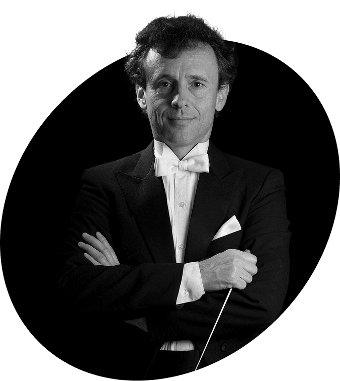 A man in a black tuxedo with a white bow tie and pocket square stands with arms crossed, holding a conductor's baton, against a dark background.