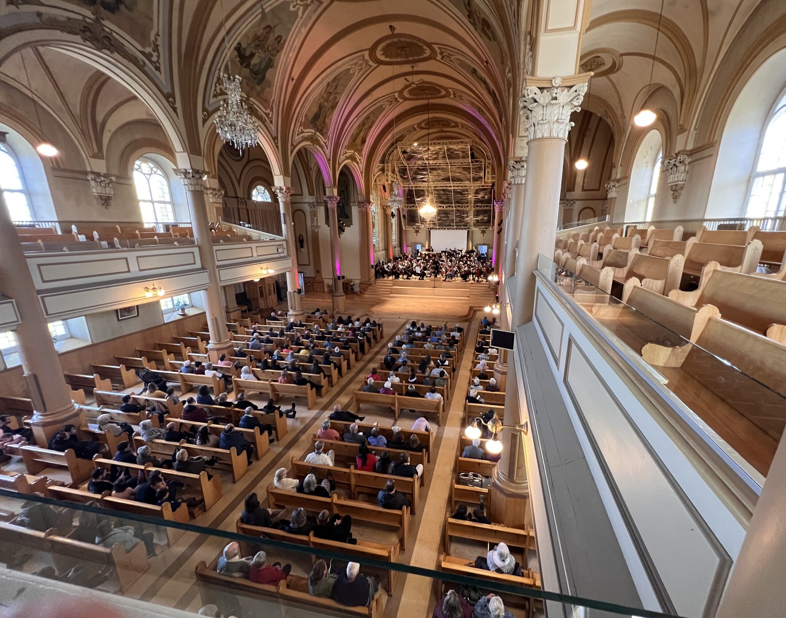 A large audience sits in wooden pews inside a grand, ornate church with high arched ceilings, watching a choir or orchestra perform at the front. Light streams through tall windows, illuminating the spacious interior.