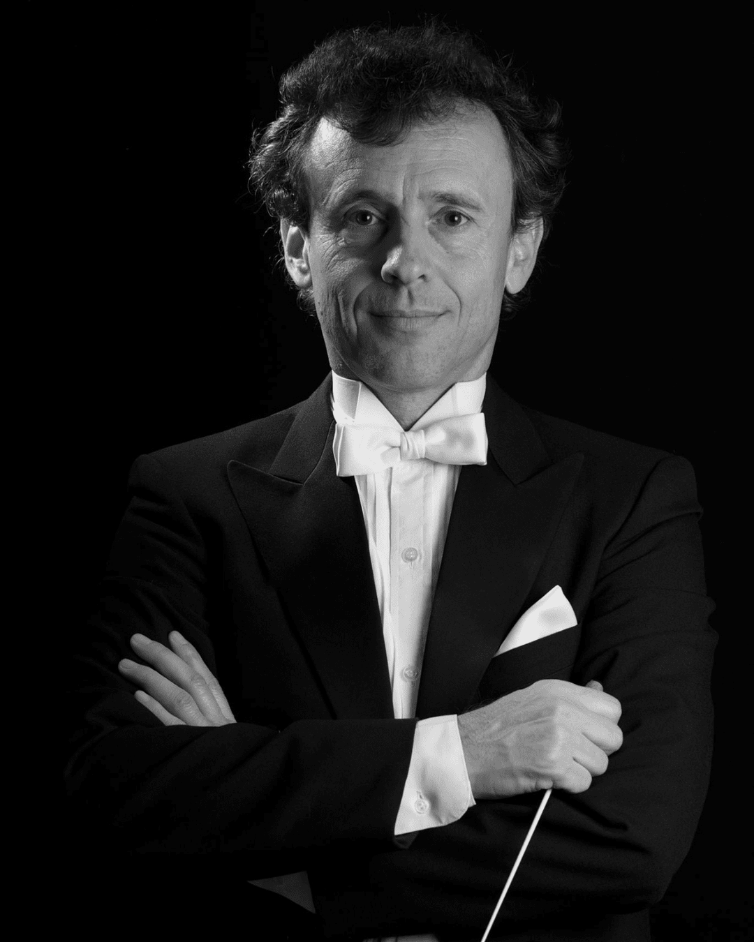 A man in a tuxedo with a white bow tie stands against a black background, arms crossed, and holding a conductor’s baton. He looks directly at the camera with a slight smile.