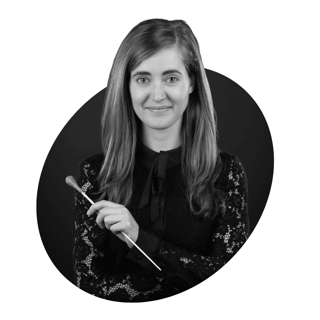 A woman with long hair wearing a black lace top smiles while holding a music conductor’s baton and a brush in front of a dark background.