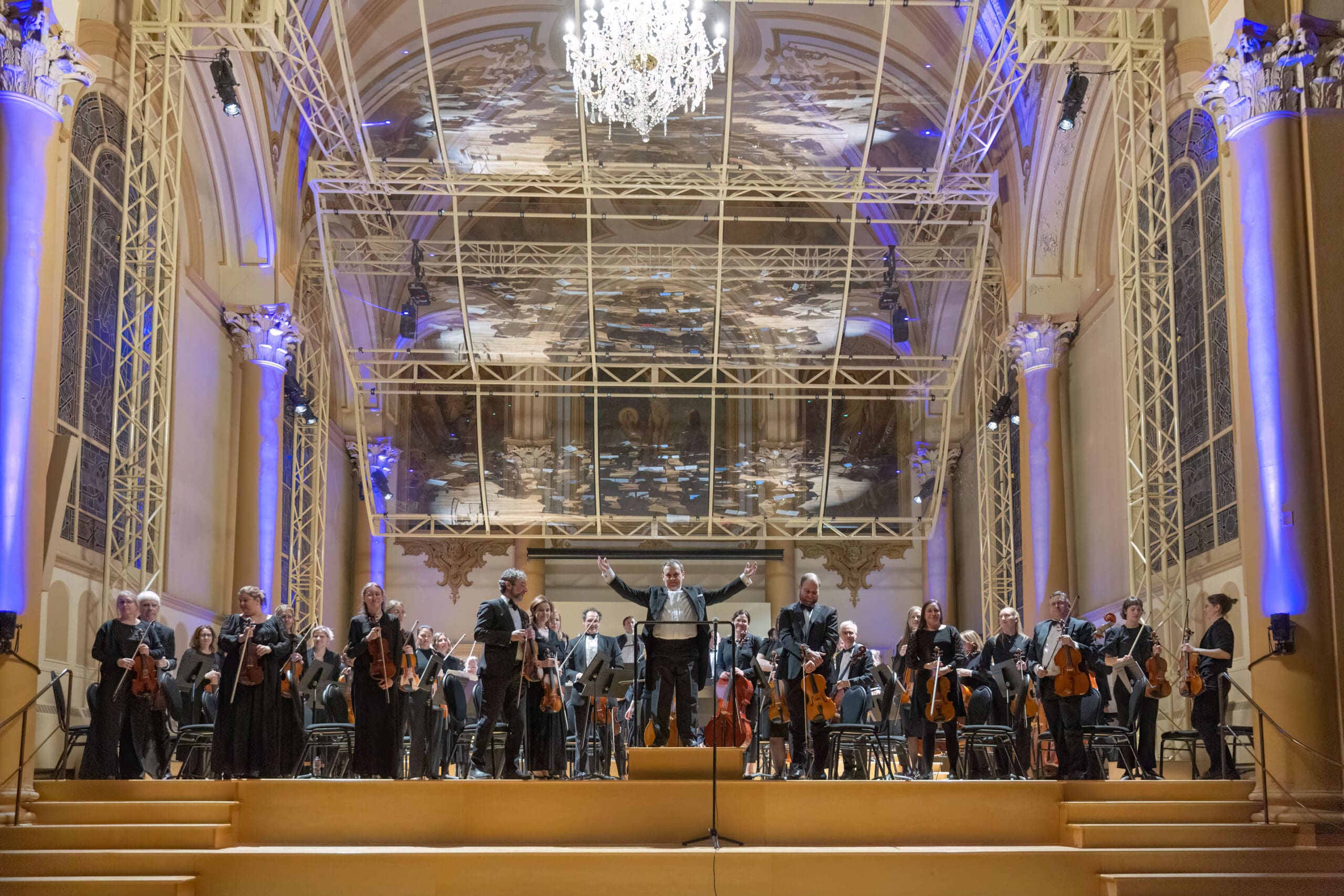 An orchestra stands on stage in a grand, ornate concert hall with high arches and a chandelier. The conductor stands at the center, facing the audience, as musicians hold their instruments and bow.