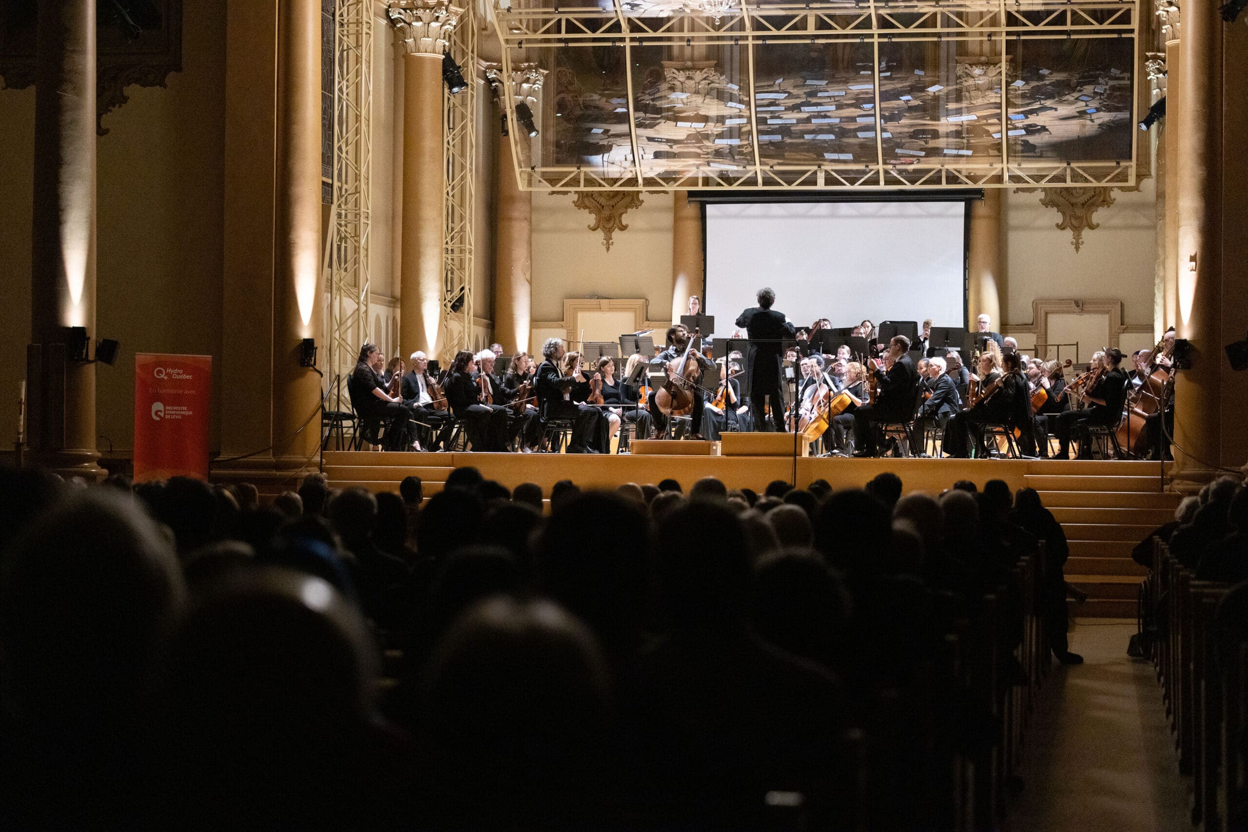 A conductor leads a full orchestra on stage in a grand concert hall, with an audience seated in the foreground. The hall is warmly lit, highlighting the musicians and their instruments.