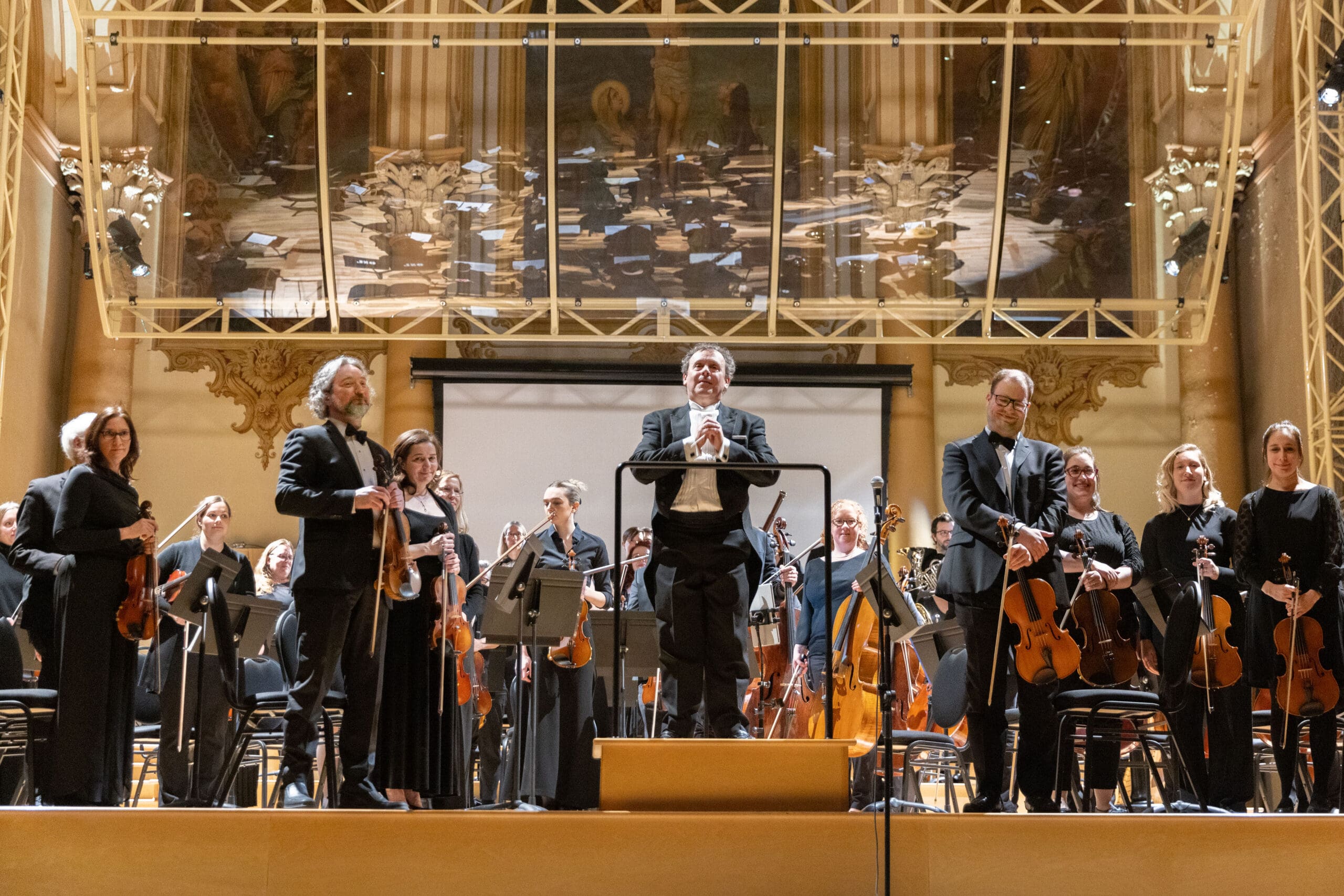 A symphony orchestra stands on stage with their conductor at the center, facing the audience. Musicians hold their instruments, and a mirrored wall above reflects the stage. The setting is elegant and formal.