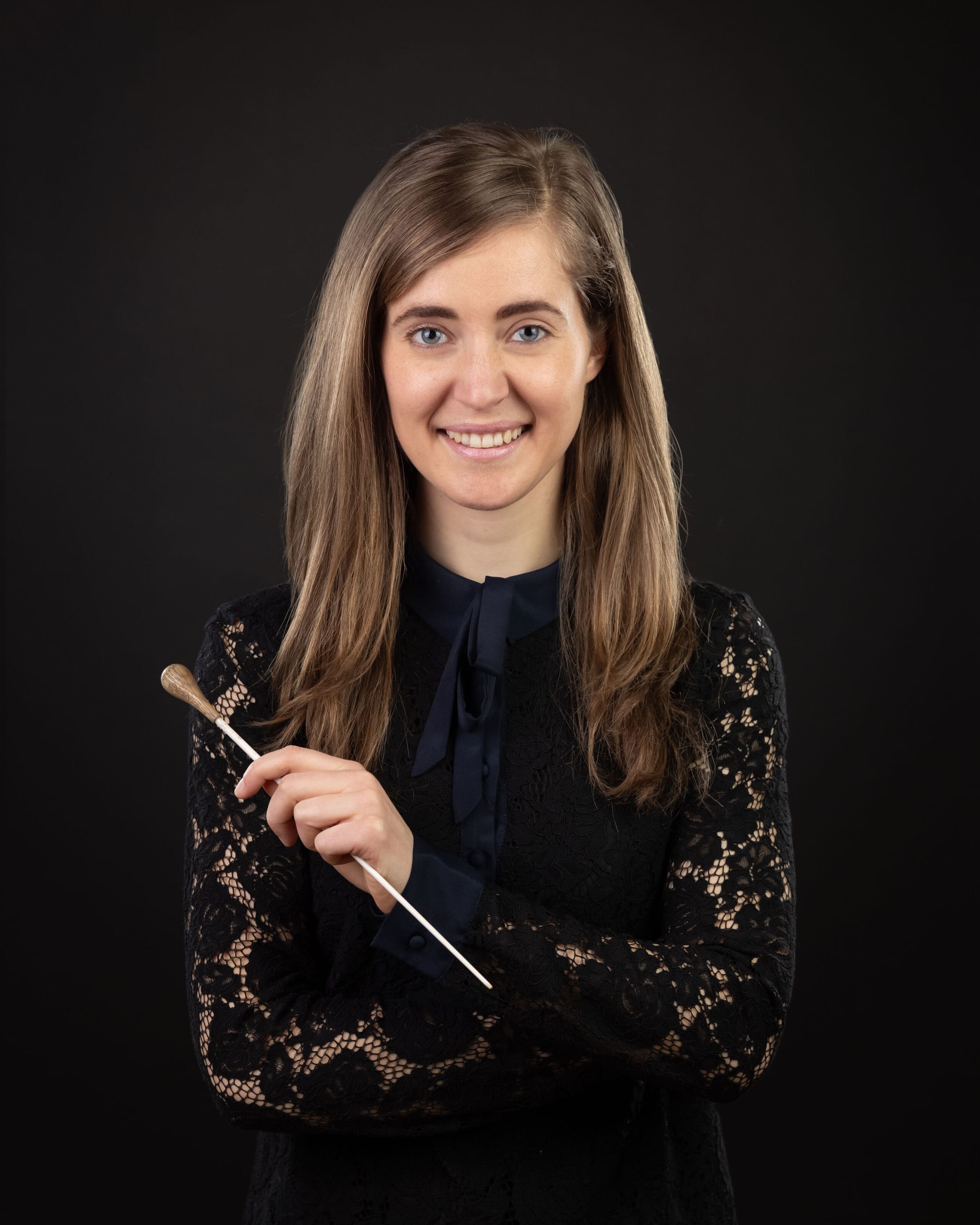 A young woman with long brown hair, wearing a black lace top, smiles and holds a conductor's baton in front of a plain dark background.