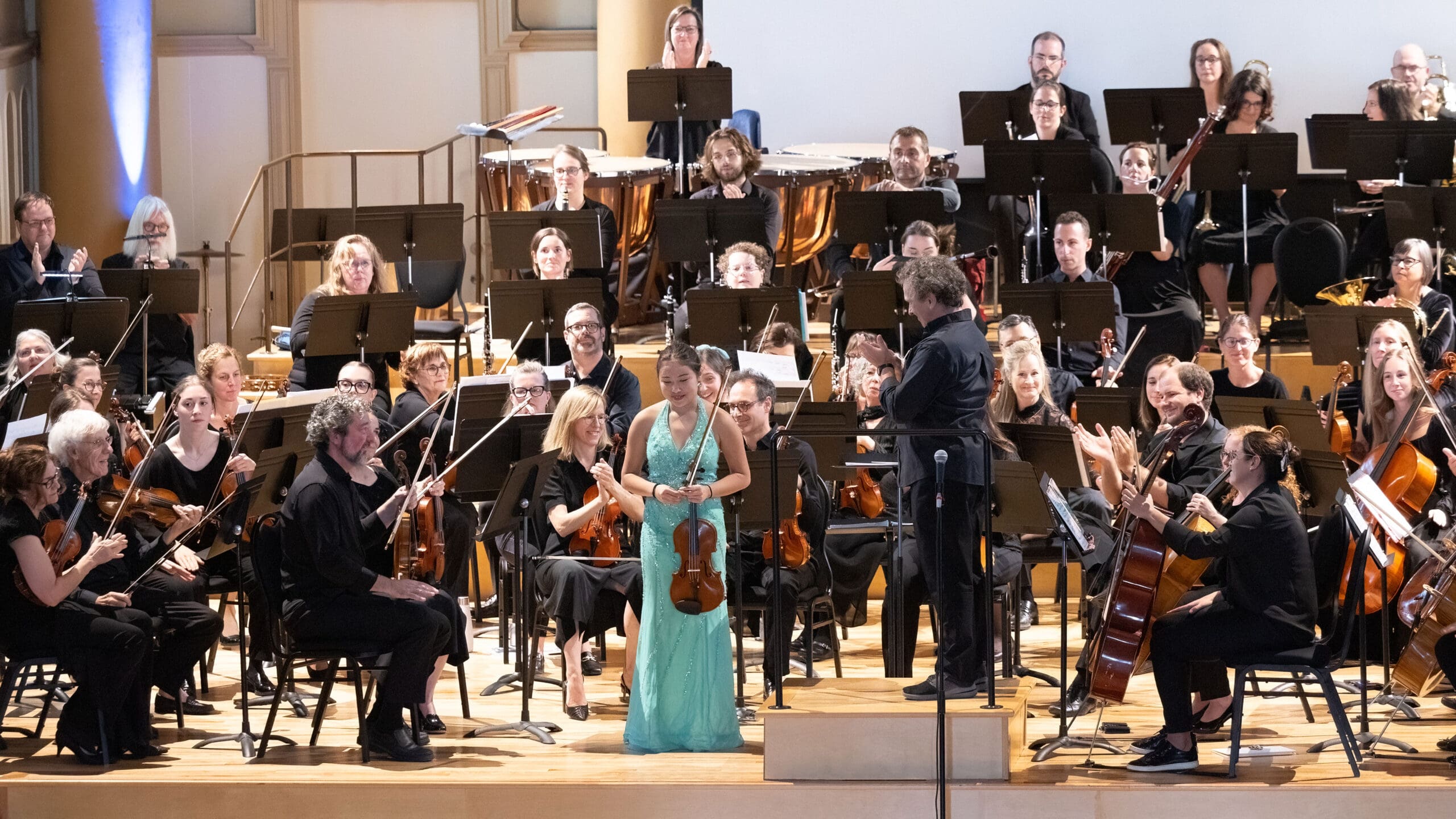 A woman in a turquoise dress stands with a violin at center stage, smiling, as an orchestra of musicians in black attire applauds her in a concert hall.