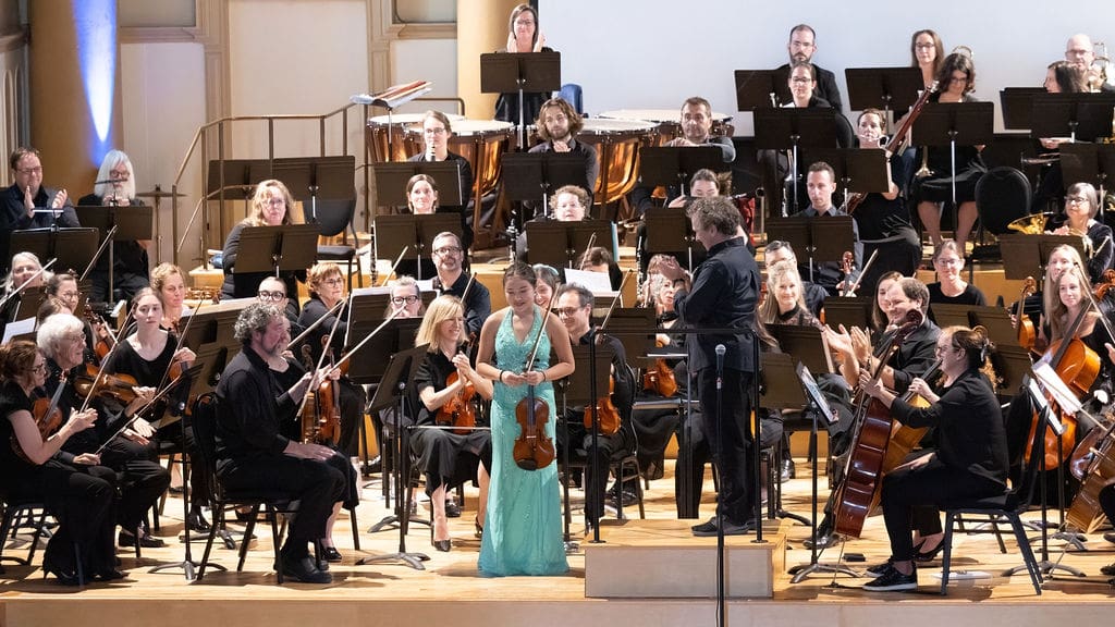 A symphony orchestra performs on stage. A young woman in a turquoise dress stands at the center holding a violin, while the conductor and musicians around her clap and smile. The stage is well-lit and full of music stands.